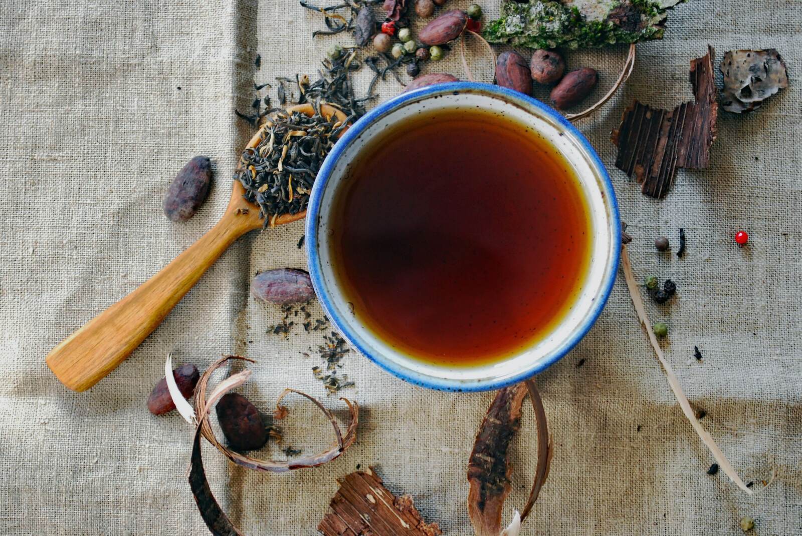 A bowl of dark brewed tea on burlap, surrounded by loose tea leaves, bark, and a wooden spoon
